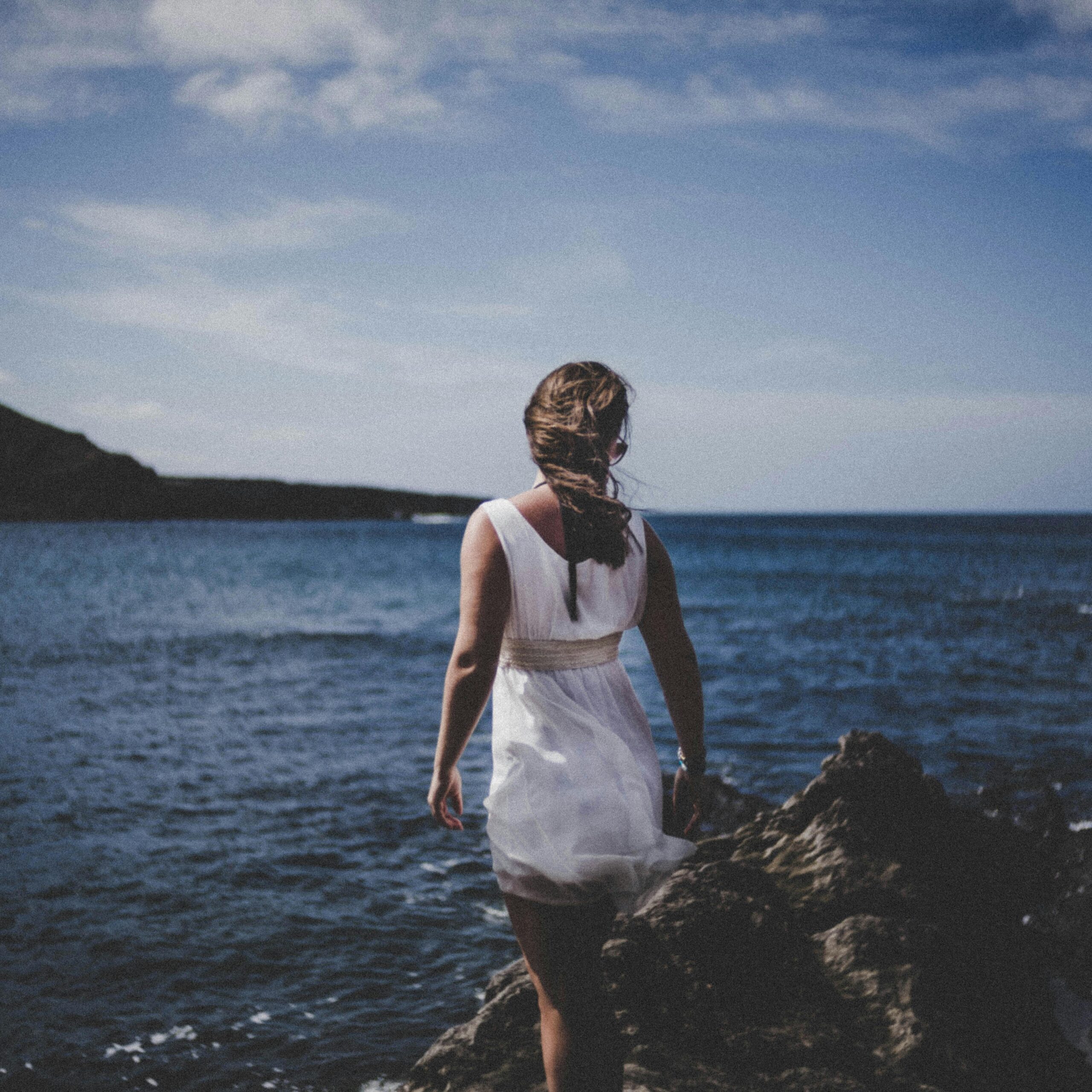 A woman in a white dress enjoying a rocky beach during a beautiful summer sunset.